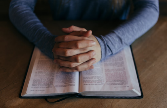 Someone with hands clasped together is sitting at a table with an open Bible or similar book. They are wearing a long-sleeved, blue-gray shirt.