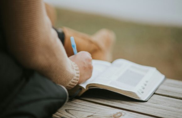 Person wearing a sweater and uniform with a Pizza Ranch logo, sitting outdoors on a wooden bench, writing in a notebook with a pen.