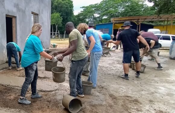 Group of people working together to pour concrete outside a building, some wearing hats and casual clothes, with trees and parked cars in the background.