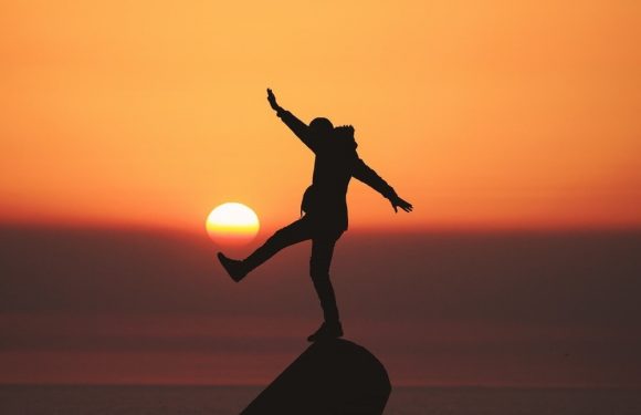 Silhouette of a person balancing on a rock at sunset, with one arm raised and the other outstretched, against an orange and pink sky with the sun near the horizon.