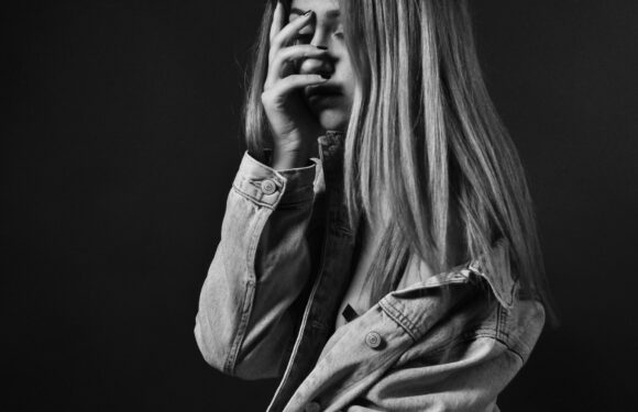 A young woman with long, straight hair is covering her face with one hand, wearing a denim jacket, against a dark background.