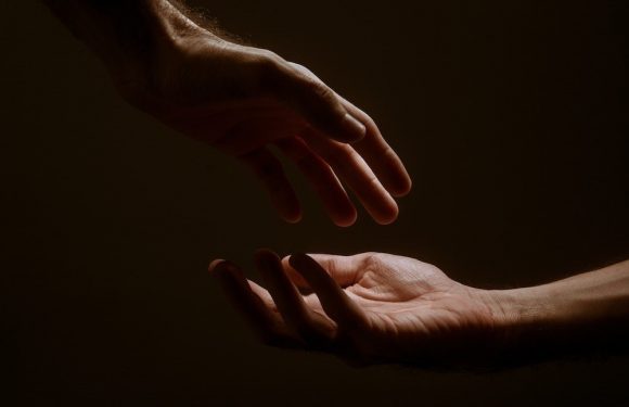 Two hands reaching toward each other in a dark background, one above and one below, with fingers almost touching in a gentle, tender gesture.