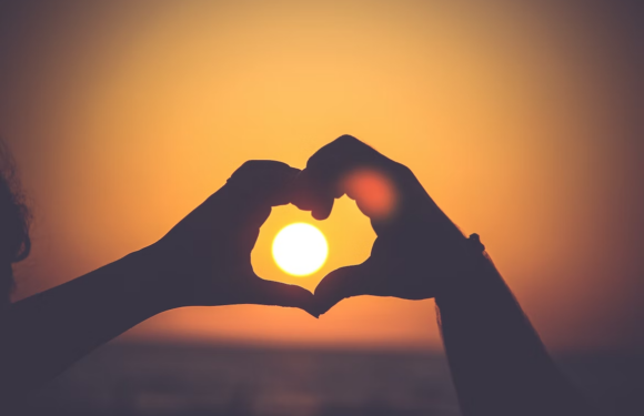 Two hands form a heart shape around the setting sun during sunset, with a warm orange sky in the background. The scene is serene and romantic.