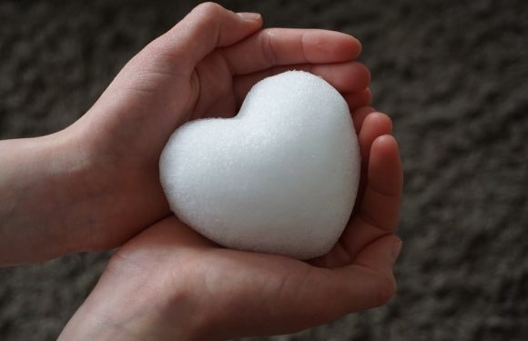 Two hands hold a white heart-shaped object made of foam or soft material against a blurred dark background.