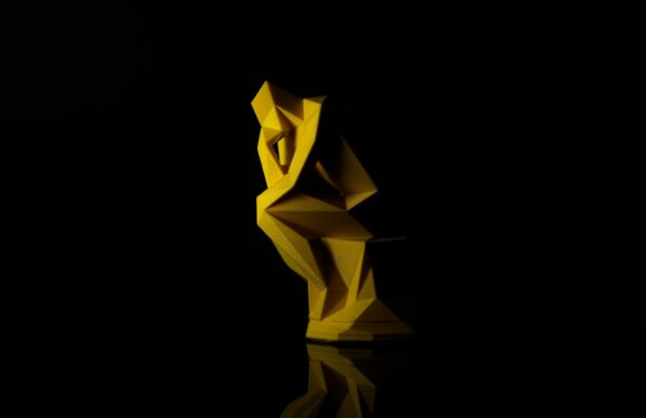 A yellow, geometric sculpture of a squirrel sitting against a black background, with its tail curled around its body and reflected on a shiny surface.