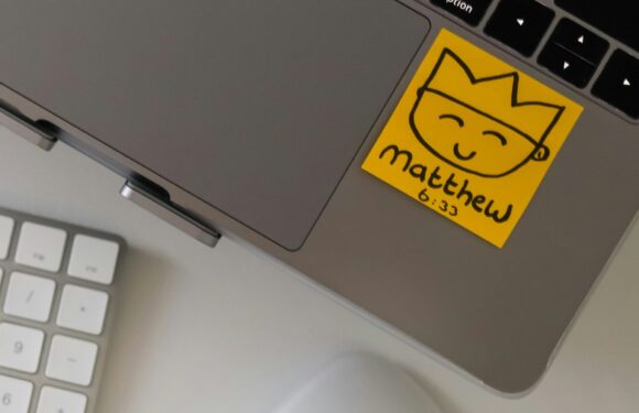 A yellow sticky note with a happy, smiling face drawing, a crown, and the name "Matthew" written below, placed on the corner of a laptop keyboard.