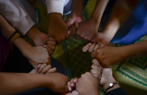 Several hands of different skin tones are joined together in a circle, symbolizing unity and teamwork. The background features a patterned carpet.