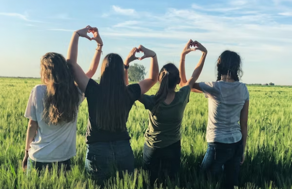 Four women stand in a green field under a blue sky, facing away, with three making heart shapes with their hands raised above their heads.