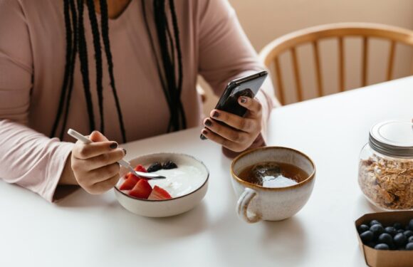 Person in a long-sleeve shirt holding a phone and spoon, sitting at a table with a bowl of yogurt topped with berries, a cup of tea, and cereal in a jar.