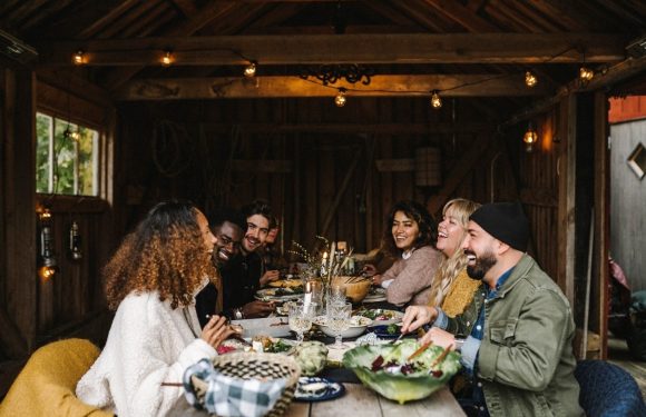 A diverse group of friends enjoying a meal together at a wooden table in a rustic setting, with some wearing casual and cozy attire.