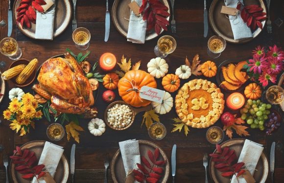 A festive Thanksgiving dinner table set with plates, glasses, silverware, and napkins, featuring a roasted turkey, pumpkins, corn, flowers, candles, and fall decorations.