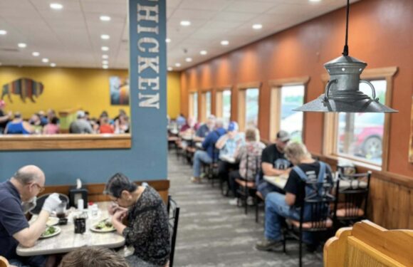People dining in a casual restaurant with warm-colored walls, wooden accents, and large windows letting in natural light. Several tables are filled with customers eating.