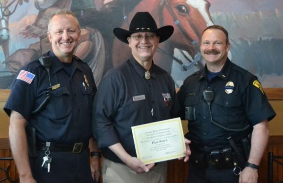 A man in Western attire holding a certificate is flanked by two police officers in uniform, standing in front of a mural of horses and riders.