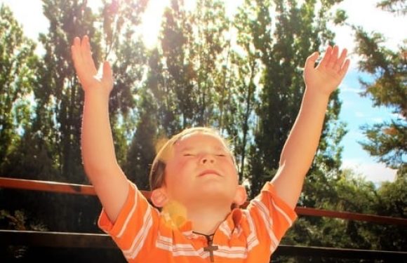 A young child in an orange striped shirt stands outdoors with arms raised towards the sky, sunlight shining through trees in the background.