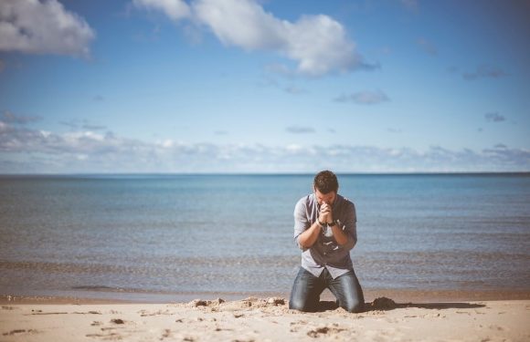 A man kneels on the sandy beach with the ocean and cloudy sky in the background, head bowed and hands clasped in prayer or reflection.