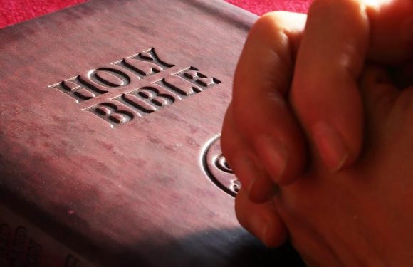 A hand is holding a pen over a Bible with "Holy Bible" embossed on the cover, placed on a red textured surface.