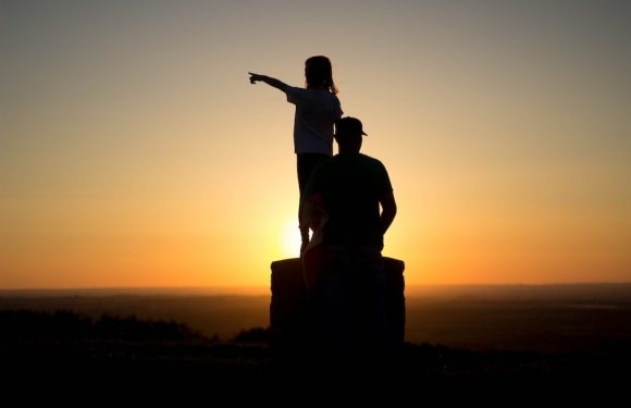 A silhouette of a woman and a child at sunset, with the woman sitting on a rock and the child standing on her lap, pointing into the distance.