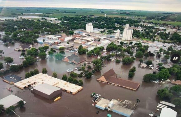 An aerial view of a town flooded with water covering streets, buildings, and trees, with some buildings partially submerged in floodwaters.