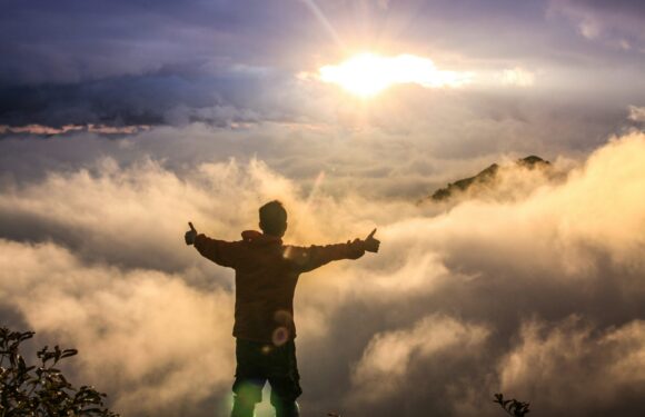 A person with arms outstretched and thumbs up standing on a mountain top at sunrise or sunset, surrounded by clouds and mist, with distant mountains in the background.
