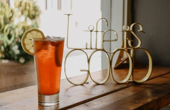 A tall glass of iced tea with a lemon wedge, placed next to a decorative "Life is good" metal sign on a wooden surface near a window.