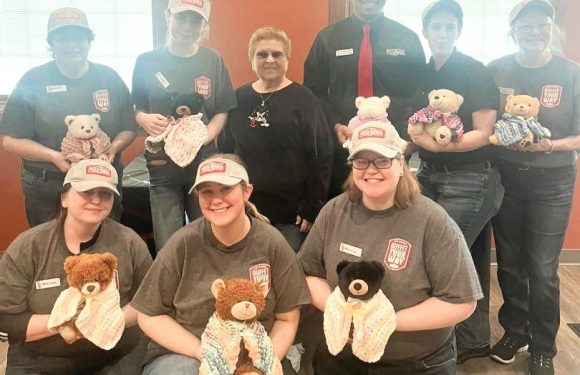 A group of eight people, some in Pizza Ranch employee uniforms holding teddy bears and handmade blankets, pose inside a room with orange and beige walls.