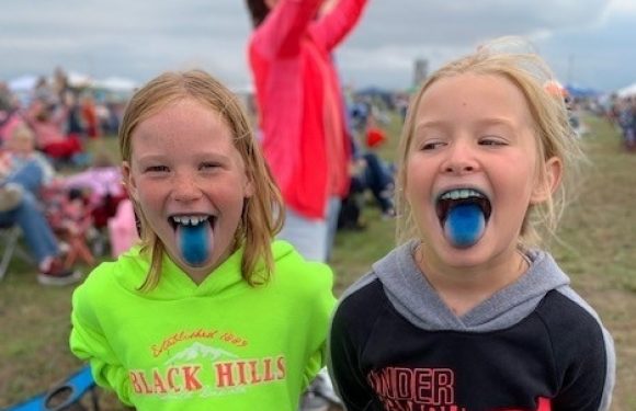 Two smiling girls with blue tongue and freckles, one in a bright green hoodie and the other in a black Under Armour hoodie, outdoors at an event with many people.