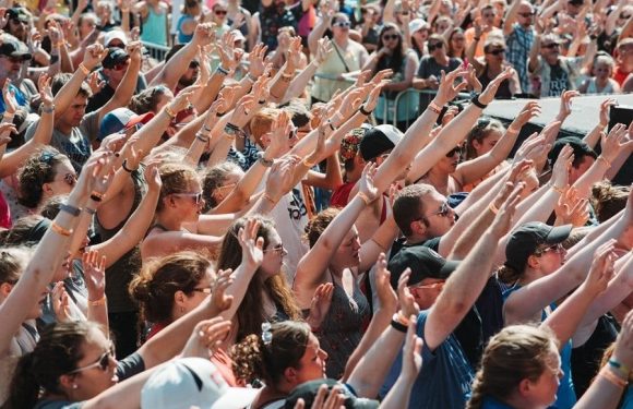 A large crowd of people outdoors, many with arms raised and hands open, participating in an event or concert in bright sunlight.