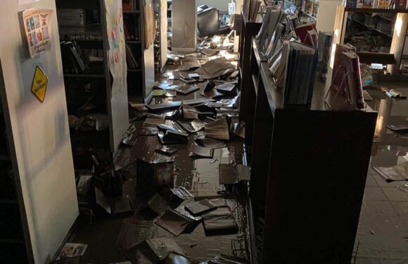 Messy library aisle with books scattered on wet floor, possibly due to flooding; bookshelves on both sides with some books dislodged.