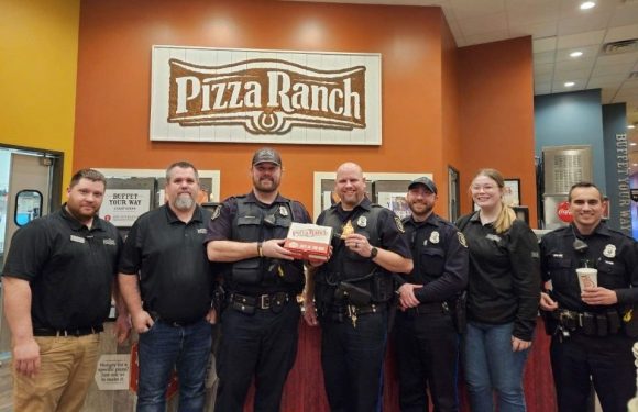 Group of seven people, including police officers and employees, standing inside a Pizza Ranch restaurant with a large sign behind them.