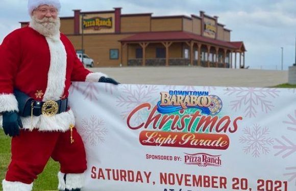 A man dressed as Santa Claus in a red suit with white trim, black boots, and a Santa hat, standing outside on grass, holding a large sign for a Christmas light parade. The background features a Pizza Ranch restaurant.