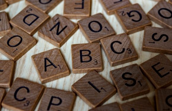 Scattered wooden Scrabble tiles with black letters and point values on a light wooden surface.