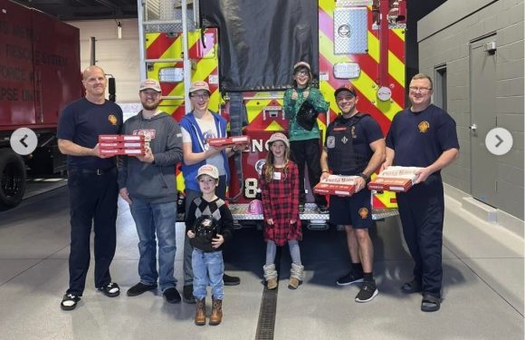 A group of people, including children, posing in front of a fire truck inside a fire station, holding pizza boxes, with some team members in uniforms.