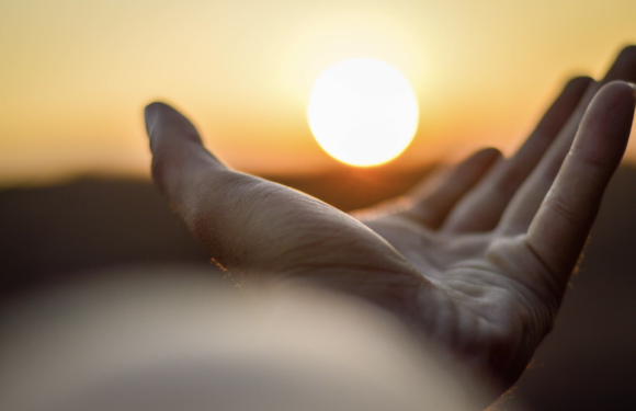 A hand reaches out toward a bright setting sun on the horizon, creating a silhouette with a warm, golden sky in the background.