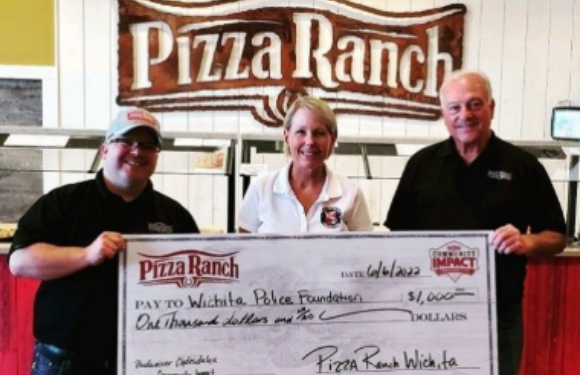 Three people stand inside a restaurant, holding a large check made out to the Wichita Police Foundation for $1,000, with a Pizza Ranch sign in the background.