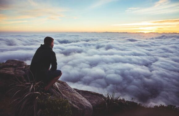A person sitting on rocks at the edge of a mountain, overlooking a vast sea of clouds during sunset, wearing a black jacket and shorts.