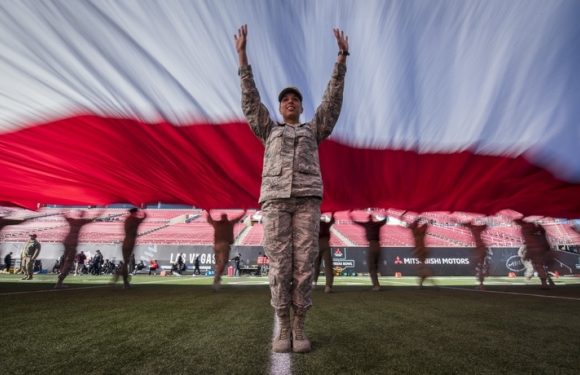 A soldier in camouflage uniform stands on a football field with arms raised, holding a large American flag that billows above. The stadium seats and other soldiers are visible in the background.