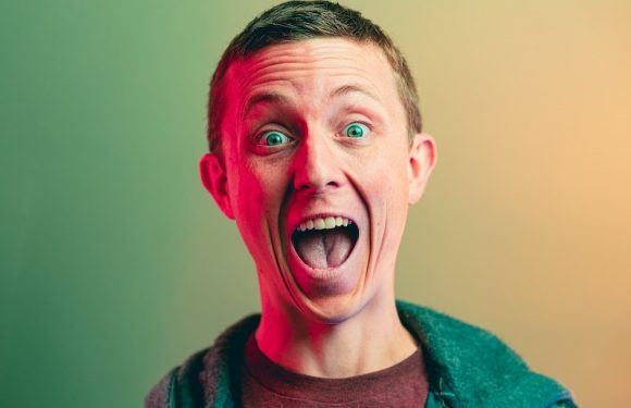 A man with short hair, wide open eyes, and mouth in an excited or surprised expression, wearing a dark hoodie over a maroon shirt, against a gradient background.