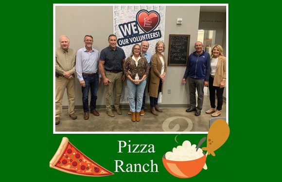 Group of nine people standing in front of a "WE ❤️ OUR VOLUNTEERS!" sign, with a pizza slice and a bowl of mashed potatoes illustration at the bottom of a green border.