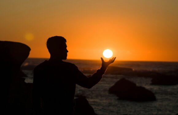 A person in a dark jacket holding a glowing ball in front of a sunset over the ocean, with rocks in the foreground, silhouette against a vibrant sky.