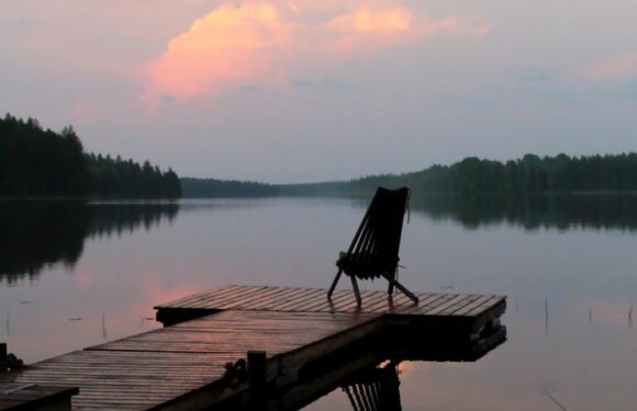 A wooden dock extends into a calm lake with a chair at the far end, surrounded by trees and a pastel-colored sky with clouds reflecting on the water.