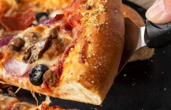 A slice of pizza with cheese, pepperoni, olives, and sausage being held with a pizza cutter on a black surface.