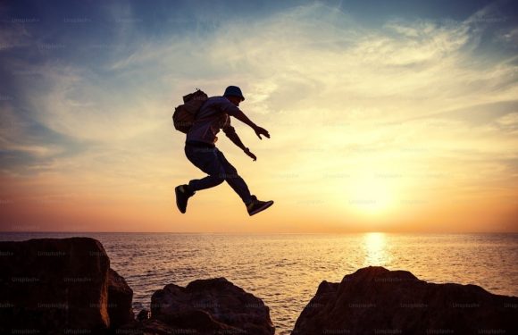Person jumping between rocks at sunset over the ocean, wearing a hat, backpack, long-sleeve shirt, jeans, and sneakers.