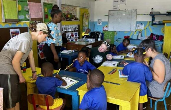 Children in blue uniforms sitting at yellow and blue desks in a classroom with teachers assisting, colorful wall decorations, and a whiteboard at the front.