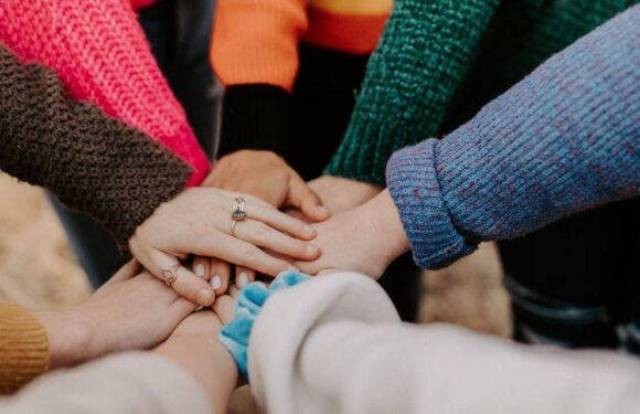 Multiple hands stacked together, some with rings, wearing colorful sweaters, signifying unity and teamwork. Some hands overlap, with a blurred background.