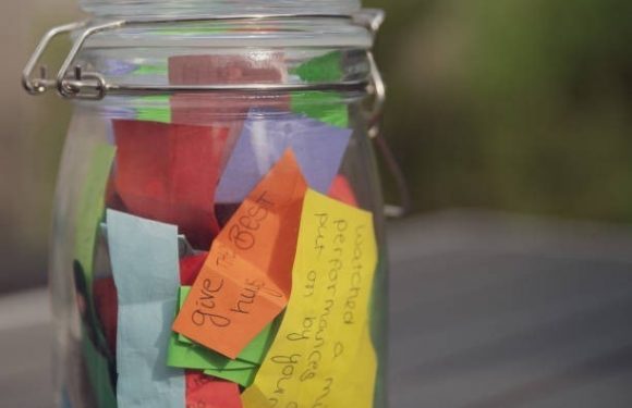A glass jar filled with colorful paper notes and handwritten messages, placed outdoors with blurred greenery in the background.