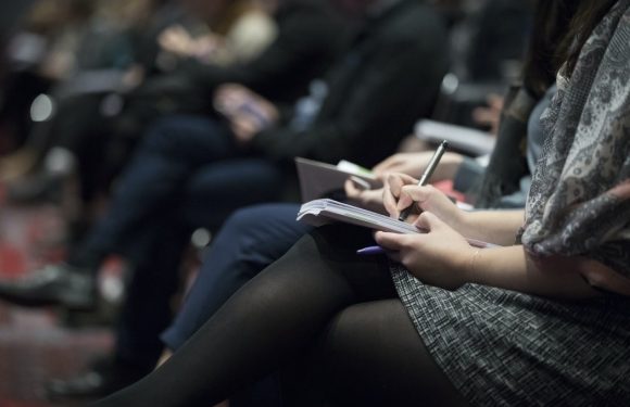 People seated in a row, taking notes or using tablets, with some blurred background attendees, in a professional or conference setting.