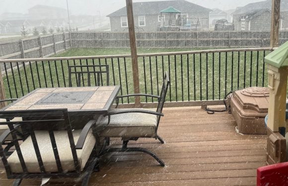 An outdoor wooden deck with a dining table and chairs under a roof, during a heavy rainstorm with visible rain and wet surfaces.