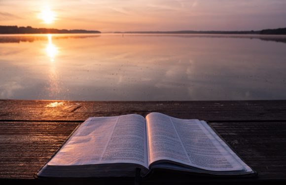 An open Bible on a wooden dock during sunrise over a calm lake with a pink and orange sky reflected in the water.