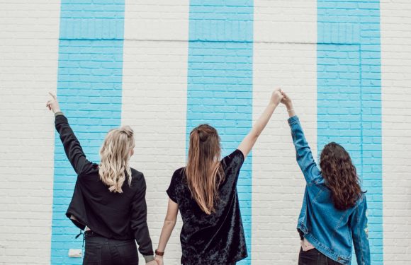 Three women standing against a white brick wall with blue vertical stripes, holding hands, and raising arms in a celebratory pose. One is wearing a black jacket, another a black top, and the third a blue jacket.