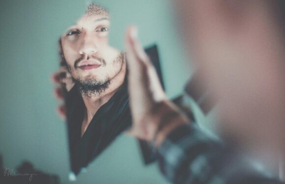 A man with facial hair and a short hairstyle is taking a selfie in front of a mirror, wearing a dark shirt, with a background that appears to be a living room.